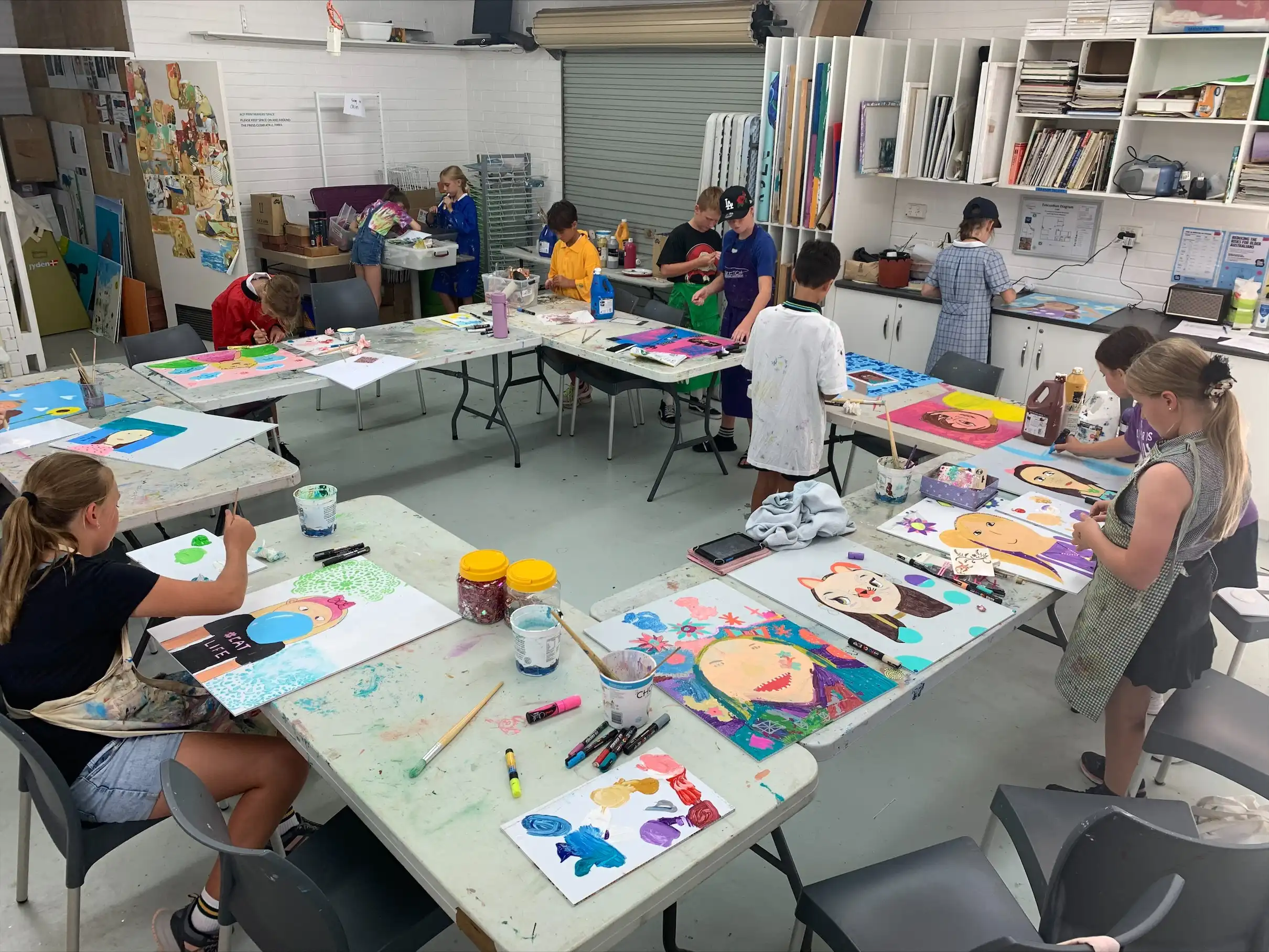 Children seated and standing around tables in an art studio, working on colourful paintings and drawings.